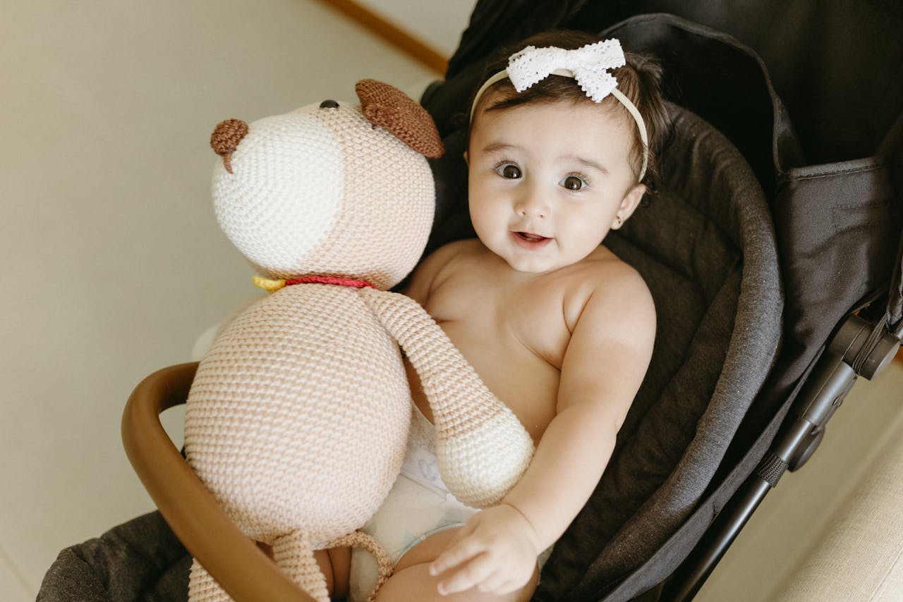 Home A cute baby in a stroller holding a knitted teddy bear inside a cozy indoor setting.